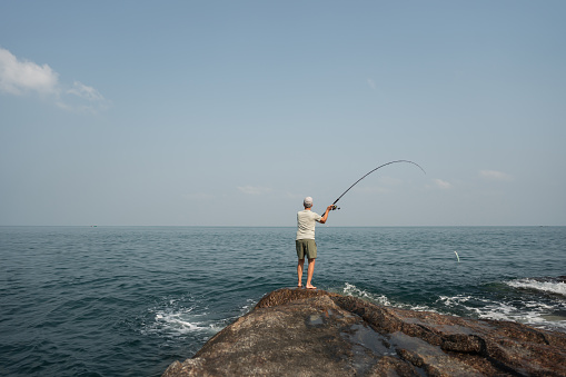 Senior fisherman casting out his fishing line into the sea from the rocky shoreline.