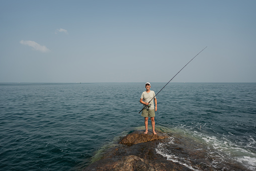 Portrait of a contented senior fisherman holding a fishing rod on the rocky shoreline.