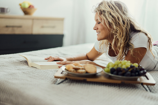 Young happy woman enjoying while reading a book during morning time on a bed.