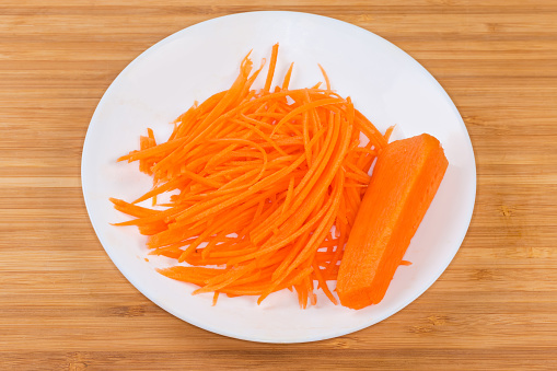 Fresh carrots chopped into thin long slices for spicy Korean carrot salad preparation on a white dish on the wooden surface
