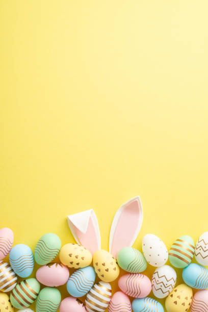 easter celebration concept. top view vertical photo of colorful easter eggs and easter bunny ears on isolated yellow background with empty space - pasen stockfoto's en -beelden