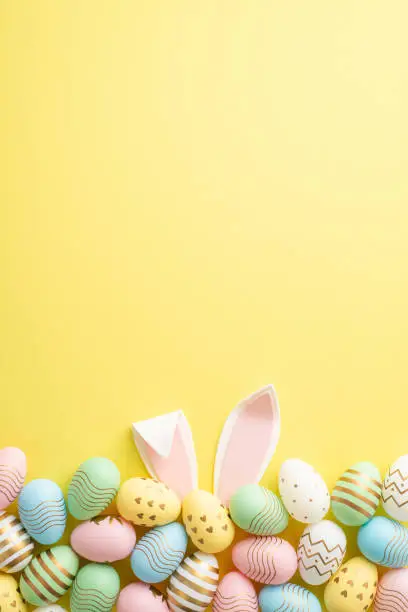 Photo of Easter celebration concept. Top view vertical photo of colorful easter eggs and easter bunny ears on isolated yellow background with empty space