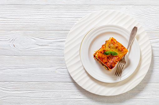 portion of Lasagna layered with ricotta cheese, ground beef, mushrooms, zucchini and tomato sauce on white plate on white wooden table, horizontal view from above, flat lay, copy space