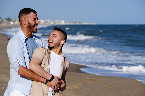 Happy male couple smiling while embracing on the beach. Lgbt concept.
