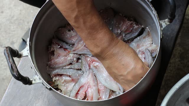 Thai woman mixing small fish with salt inside a stainless steel pot.