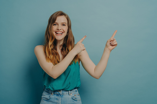 Young positive excited woman pointing to right side with index fingers of both hands while advertising product or service looking at camera broadly smiling isolated on blue background with free space