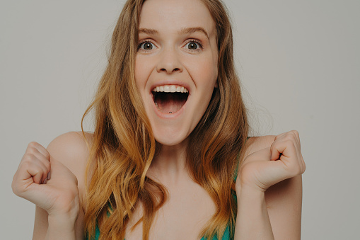 Excited amazed young woman with natural makeup and slightly wavy hair clenching fits, screaming from excitement, looking in camera with open mouth and surprised expression, isolated on grey background