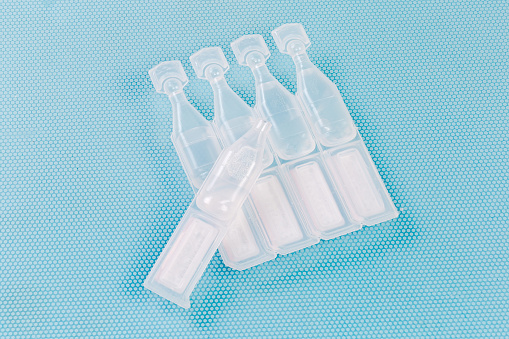 Eye drops in small disposable plastic ampoules connected in block and one open ampoule separately, close-up on a blue surface