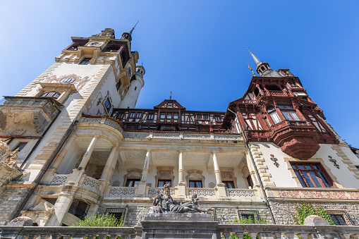 Sinaia - Romania, July 15, 2022. Well-preserved facade of Peles Castle with fachwerk elements, large clock tower with balcony, smaller spired tower and statues of Greek god Dionysius in the foreground