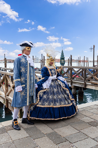 Venice - Italy. February 5, 2023: A couple of participants in elegant baroque costumes pose on the embankment at the Venice Carnival (vertical photo)