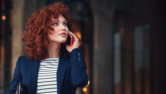 Beautiful red head business woman using smartphone talking on mobile phone in city at sunset