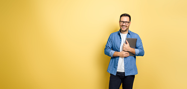 Portrait of cheerful young adult man holding laptop and standing against yellow background. Happy male freelancer looks at camera wearing eyeglasses.