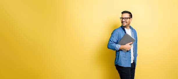 hombre sosteniendo una computadora portátil mirando por encima del hombro contra fondo amarillo - mirar hacia el otro lado fotografías e imágenes de stock