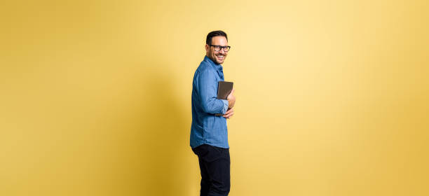 istock Handsome man with laptop standing over yellow background looking back over the shoulder. 1471614838