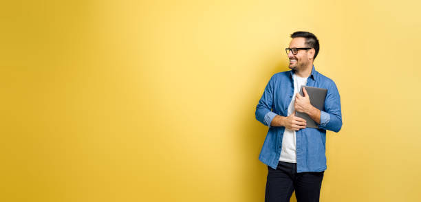 Man holding laptop looking away smiling against yellow background Smiling man holding laptop and looking away. Positive freelancer standing against yellow background. He is wearing blue denim and glasses. sideways glance stock pictures, royalty-free photos & images