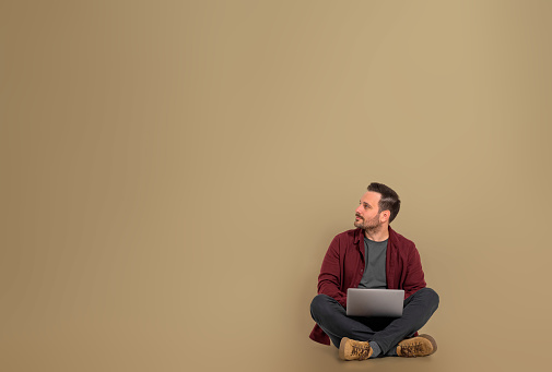 Handsome male freelancer entrepreneur working on laptop. Man sitting on brown background with crossed legs. He is looking aside at copy space.