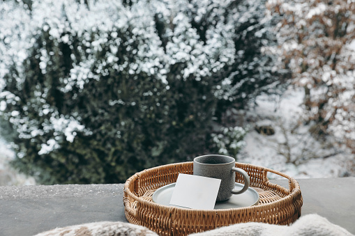 Winter windowsill still life. Cup hot coffee, tea on wicker tray. Brown wool blanket. Morning cozy breakfast concept, blurred garden, woodland background with snow covered dark evergreen trees.