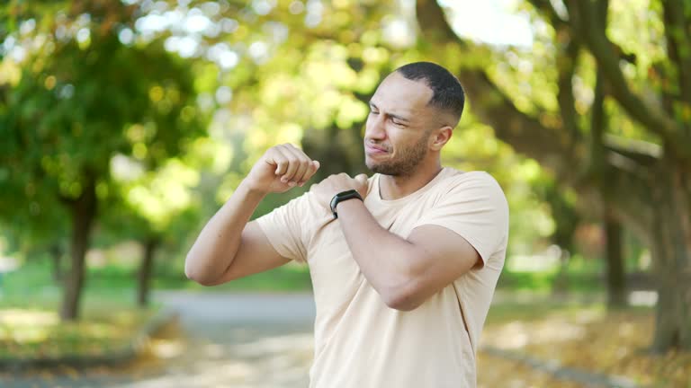 A young athletic man feels sharp pain in his shoulder during morning exercise in a city park. A sad man in a T-shirt was injured.