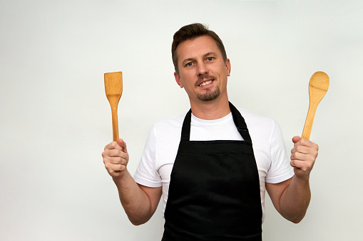 Young chef with wooden spoon and spatula isolated on white background with copy space. Young cook man in a black apron holding kitchen utensils