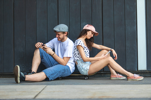Beautiful young couple sitting on skateboard and checking social media on their smart phones