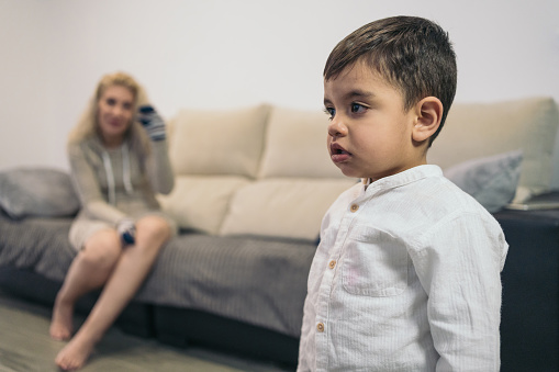 Young child with special needs because he is a person with a disability distracted by the TV ignoring his middle-aged mother who tries to play with him with a sock in her hand.