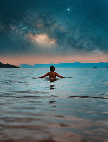 back view beautiful young woman submerged in water up to her torso relaxed with open arms and milky way in the background
