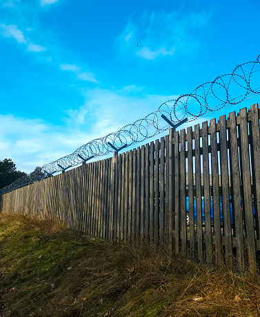 This razorwire is on top of a wooden fence that is placed atop a grass hill, with a blue sky background. Sending a strong signal of low tech security.