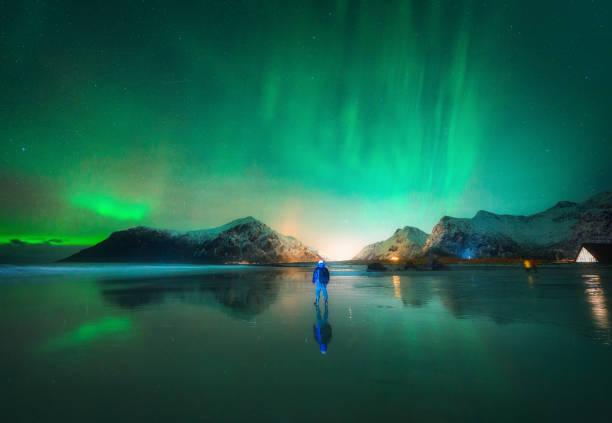 Northern Lights and man on the beach at night. Lofoten islands, Norway. Beautiful Aurora borealis. Polar lights. Winter landscape with aurora, guy, sea, starry sky, reflection in water, mountains Northern Lights and man on the beach at night. Lofoten islands, Norway. Beautiful Aurora borealis. Polar lights. Winter landscape with aurora, guy, sea, starry sky, reflection in water, mountains norway aurora borealis aurora polaris fjord stock pictures, royalty-free photos & images