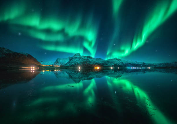 Northern lights over the snowy mountains, sea, reflection in water at night in Lofoten, Norway. Aurora borealis and snow covered rocks. Winter landscape with polar lights, city lights, sky with stars stock photo