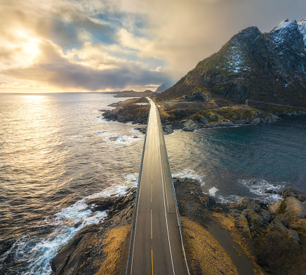 Aerial view of bridge, sea with waves and mountains at sunset in Lofoten Islands, Norway. Landscape with beautiful road, water, rocks, blue sky with clouds and golden sunlight. Top view from drone