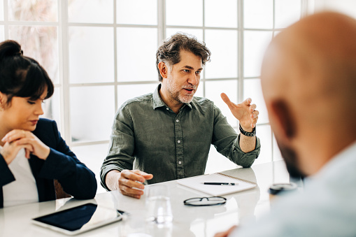 Senior man having a discussion with his team in an office. Mature business professional talking to his colleagues during a meeting. Group of diverse business people planning a project.