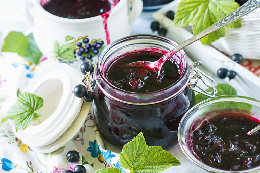 Cooked homemade black currant jam in jar with spoon on white wooden table and vivid tablecloth background, fresh black currant jam close up, food and healthy eating concept