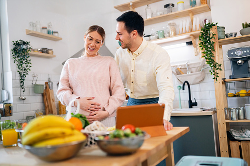Pregnant couple having a video call via digital tablet, while preparing food in the kitchen