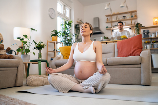 A young Caucasian pregnant woman meditating, while her husband is in kitchen
