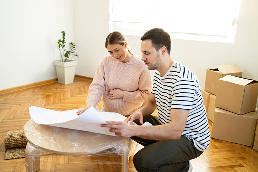 The young Caucasian couple, a man and a pregnant woman, looking at the blueprint of their new home, while making plans for decoration