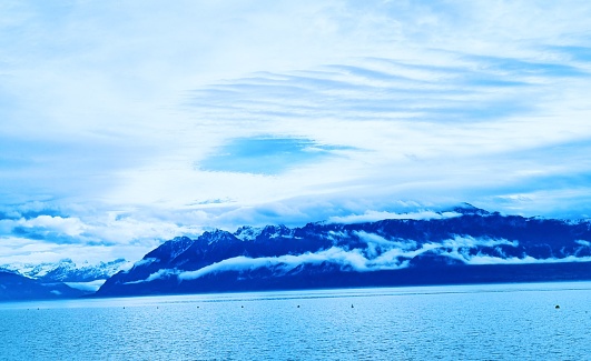 The image captures a stunning panorama of a mountainous landscape with snow-capped peaks, a pristine lake, and a sky filled with clouds. The mountains rise majestically in the background, their jagged peaks dusted with snow, and their slopes dotted with evergreen trees. The lake glistens in the foreground, reflecting the surrounding landscape with crystal clarity. Wispy clouds drift across the sky, adding a sense of movement and depth to the scene. The overall effect is one of serene natural beauty, showcasing the raw power and breathtaking vistas of the mountain landscape.