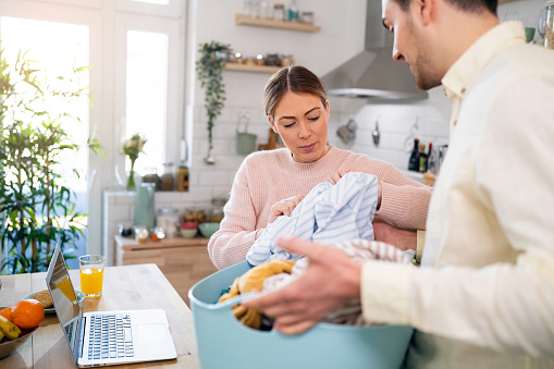 Pregnant Caucasian woman working on the laptop in the kitchen, while her husband doing the laundry