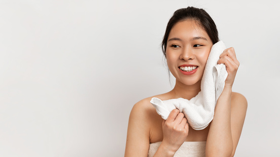 Happy beautiful asian lady drying face with soft towel looking aside, posing over white studio background, panorama with free space. Facial skincare and pampering, beauty routine concept