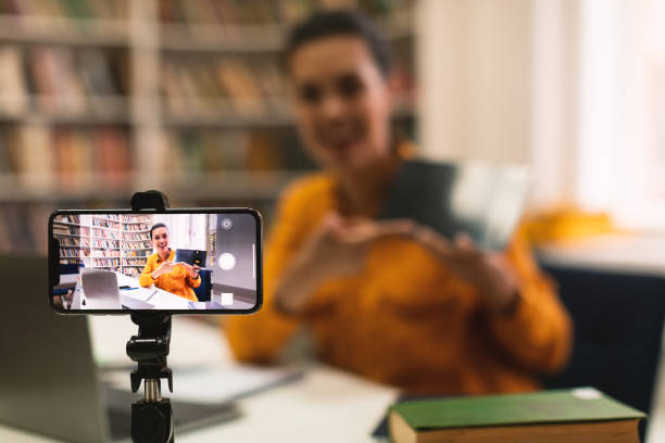 istock Female teacher shooting online lecture to students using cellphone on tripod and showing tablet, selective focus 1471651446