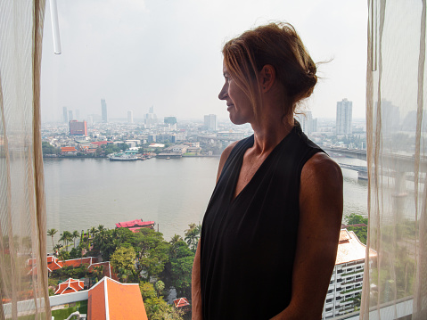 Side view of a happy mature woman standing at the window looking outside to the Chao Phraya River at Bangkok, Thailand - stay at home concept