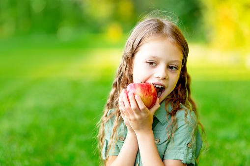 happy baby girl in summer on the lawn bites a red apple with healthy teeth on the grass and smiles, space for text