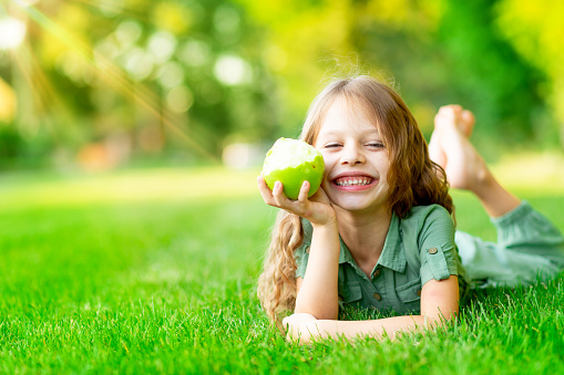 happy baby girl in summer on the lawn bites a green apple with healthy teeth on the grass and smiles, space for text