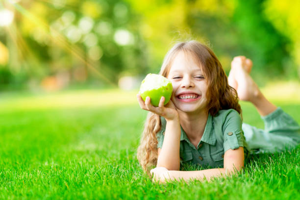 szczęśliwa dziewczynka latem na trawniku gryzie zielone jabłko ze zdrowymi zębami na trawie i uśmiechami, miejsce na tekst - apple eating little girls green zdjęcia i obrazy z banku zdjęć