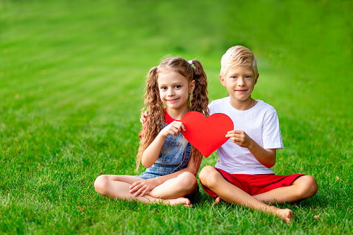 children a girl and a boy blonde with a big red heart in the summer on the lawn on the green grass having fun and rejoicing, the concept of valentine's day, space for text