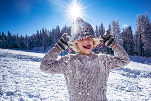 Boy sticking out tongue and covering his face whit winter hat during day on the mountain