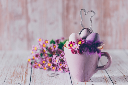 Cozy purple coffee cup with an Easter bunny cookie cutter, daisies, feathers and purple Easter eggs on wooden background. Creative color editing with added grain. Very soft and selective focus. Part of a series.