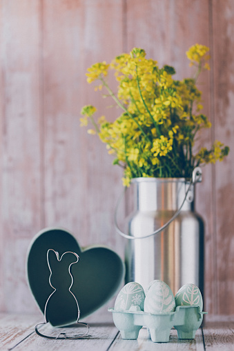 Beautiful still life of self painted mint green Easter eggs, an Easter bunny cookie cutter and a silver colored milk pot as a vase with a oilseed rape bouquet in front of a box with a heart shape. Creative color editing with added grain. Very soft and selective focus. Part of a series.