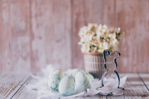 Beautiful self painted mint green Easter eggs and white feathers, an Easter bunny cookie cutter and a pot with white flowers on wooden background background. Creative color editing with added grain. Very selective and soft focus. Part of a series.