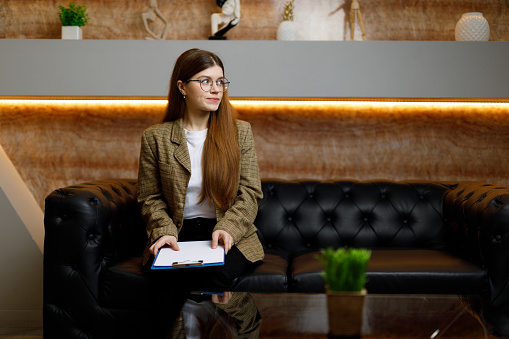 A young woman in a jacket is waiting for an interview, sitting on a sofa in the lobby with a resume.