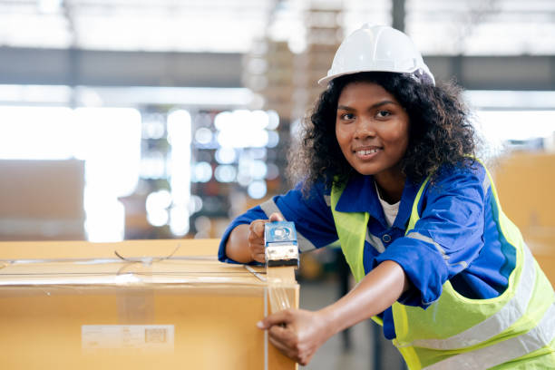 istock Young beautiful female worker wearing Safety helmet using tape dispenser gun while packing cardboard boxes for the shipment in a distribution warehouse. 1471669965